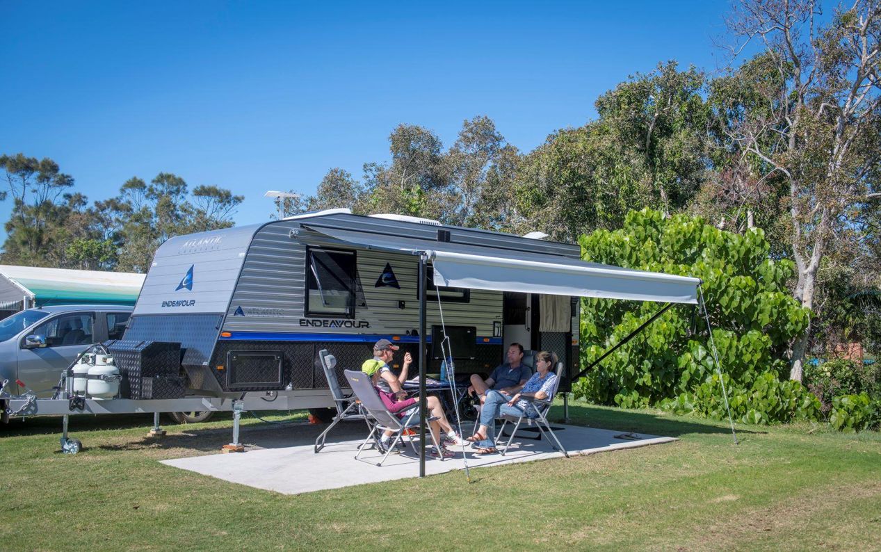 Caravan with its awning extended, parked on a concrete slab site at Dicky Beach Holiday Park, with a group of guests relaxing at an outdoor table underneath.