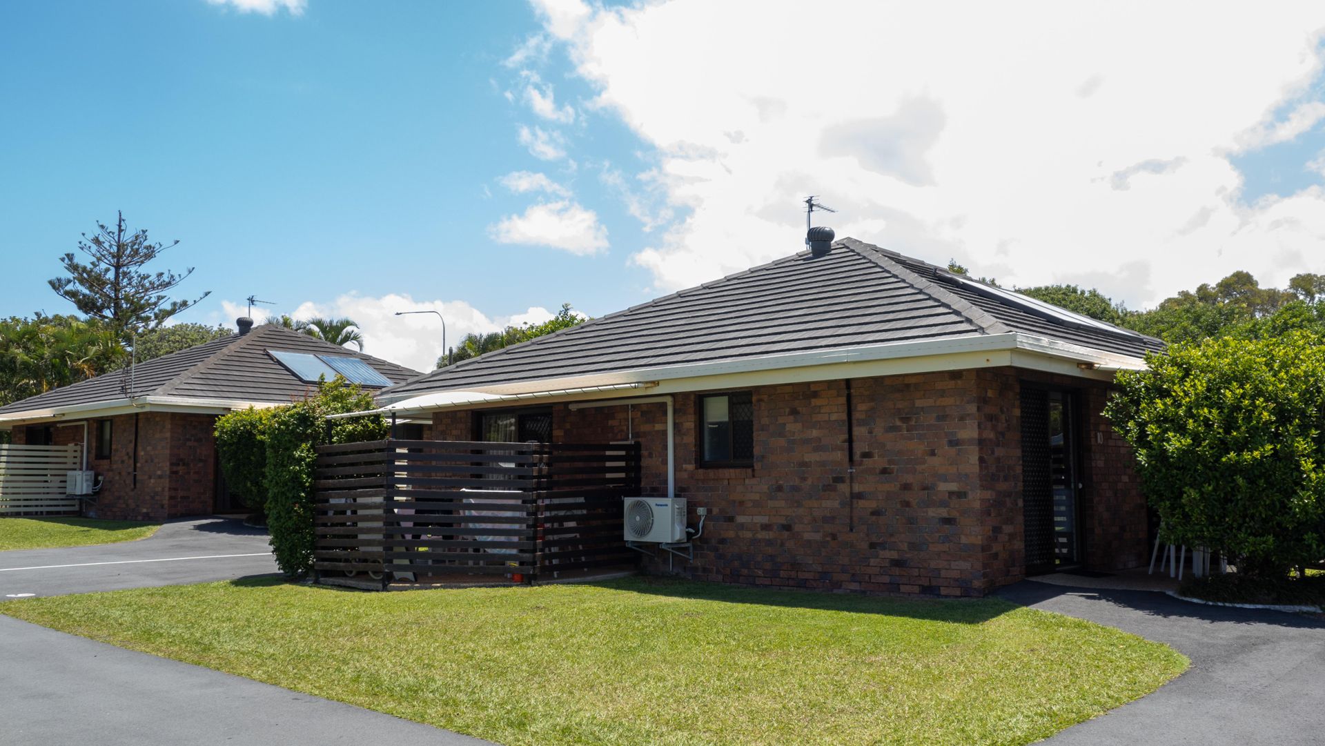 Exterior of a brick studio unit at Dicky Beach Holiday Park, with a tiled roof, solar hot water panel, and a small private courtyard screened by timber slats.