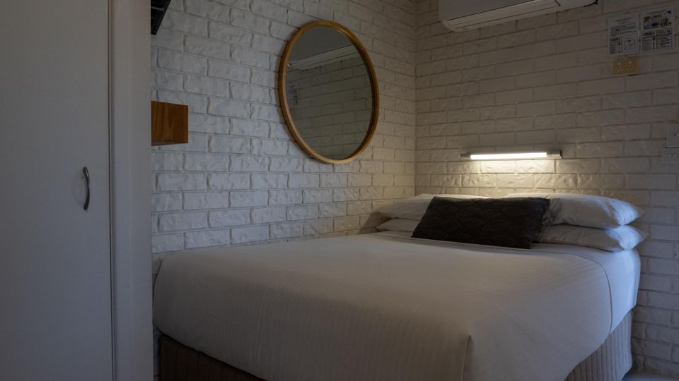 Studio unit bedroom at Dicky Beach Holiday Park with a made-up queen bed, round mirror, wall-mounted air conditioning and a reading light.
