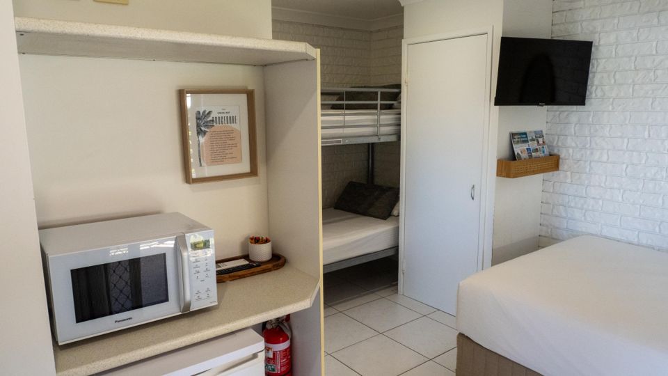 Studio unit interior at Dicky Beach Holiday Park showing the kitchenette with a microwave, a wall-mounted TV, and a set of bunk beds visible through the doorway.